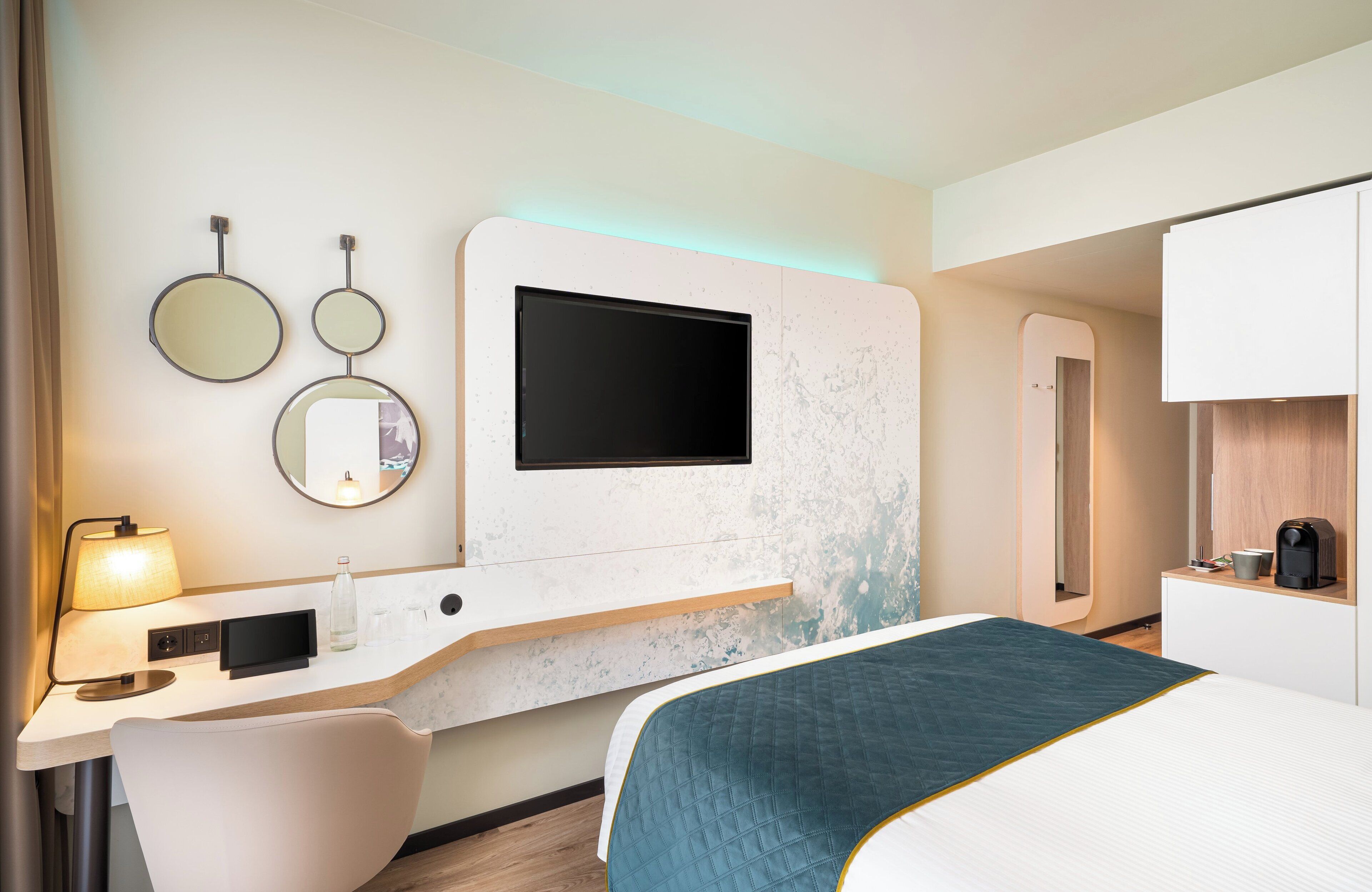 Ein modernes Hotelzimmer mit Bett, Schreibtisch mit Lampe, Flachbildfernseher und Kaffeemaschine.