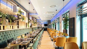 A long dining area with multiple tables and chairs, a high ceiling with recessed lighting, and potted plants along the walls. - Leonardo Hotel Hamburg Altona (Hamburg)
