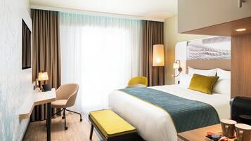 A hotel room with a bed, a desk, a chair, a small table, and a window with curtains.
