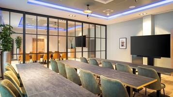Un salón de conferencias con mesas largas y sillas, un televisor de pantalla plana grande, e iluminación empotrada y motivos decorativos en el techo.