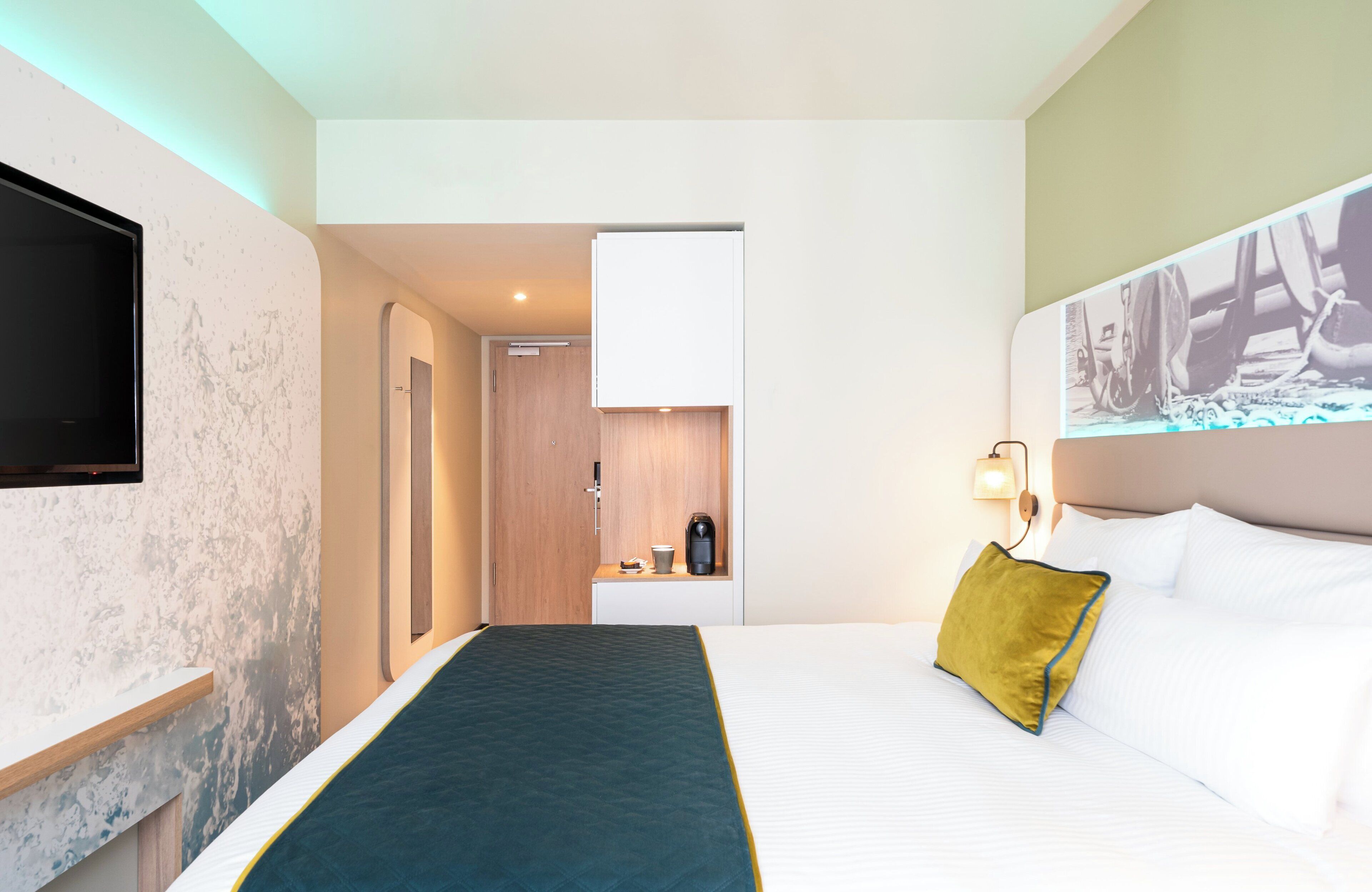 A hotel room with a bed, a TV mounted on the wall, a coffee machine, and a painting on the headboard.
