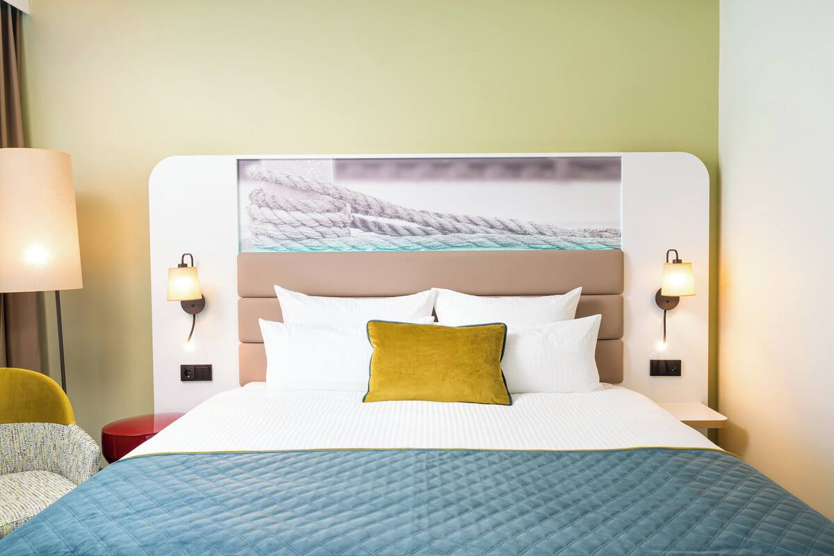 Habitación de hotel con una cama grande, dos lámparas de noche, una silla amarilla y una obra de arte colgada en la pared.
