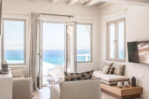 A modern living room with a sofa, chairs, a coffee table, and a TV. Large windows offer a view of the sea.