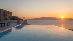 Una piscina a sfioro con parete in pietra e sedie a sdraio, affacciata su un mare calmo al tramonto.