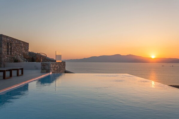 An infinity pool with a stone wall and lounge chairs, overlooking a calm sea at sunset.