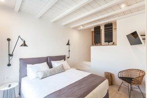 A bedroom with a large bed, two wall-mounted lamps, a small table, a chair, and a window with blinds.