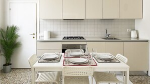 Une cuisine de taille réduite avec des meubles blancs, une cuisinière à gaz et un évier. On y trouve une table à manger mise avec des assiettes, des verres et des couverts. Une plante en pot est visible sur le côté gauche de la cuisine.