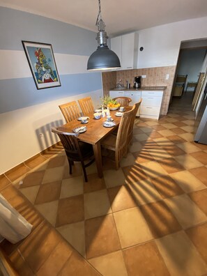 Un comedor con mesa y sillas de madera, una cocina al fondo y suelo de baldosas.