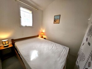 Ein Einzelbett mit Holz-Kopfstück, eine Nachttischlampe, ein Fenster mit Jalousien und ein gerahmtes Bild an der Wand.