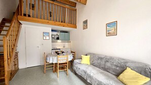A compact living space with a wooden staircase, a kitchenette, a dining area with a table and chairs, and a grey sofa with a yellow pillow.