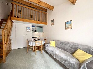 Un espace de vie compact comprenant un escalier en bois, un coin cuisine, un espace repas avec une table et des chaises, et un canapé gris agrémenté d’un coussin jaune.