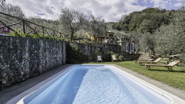 Una piscina con un muro de piedra en un lado, una terraza de madera con sillones de descanso, y una casa al fondo.