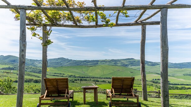 A wooden pergola with two wooden chairs and a small table, overlooking a green valley with hills. - Podere Val D'Orcia - Tuscany Equestrian (Sarteano)