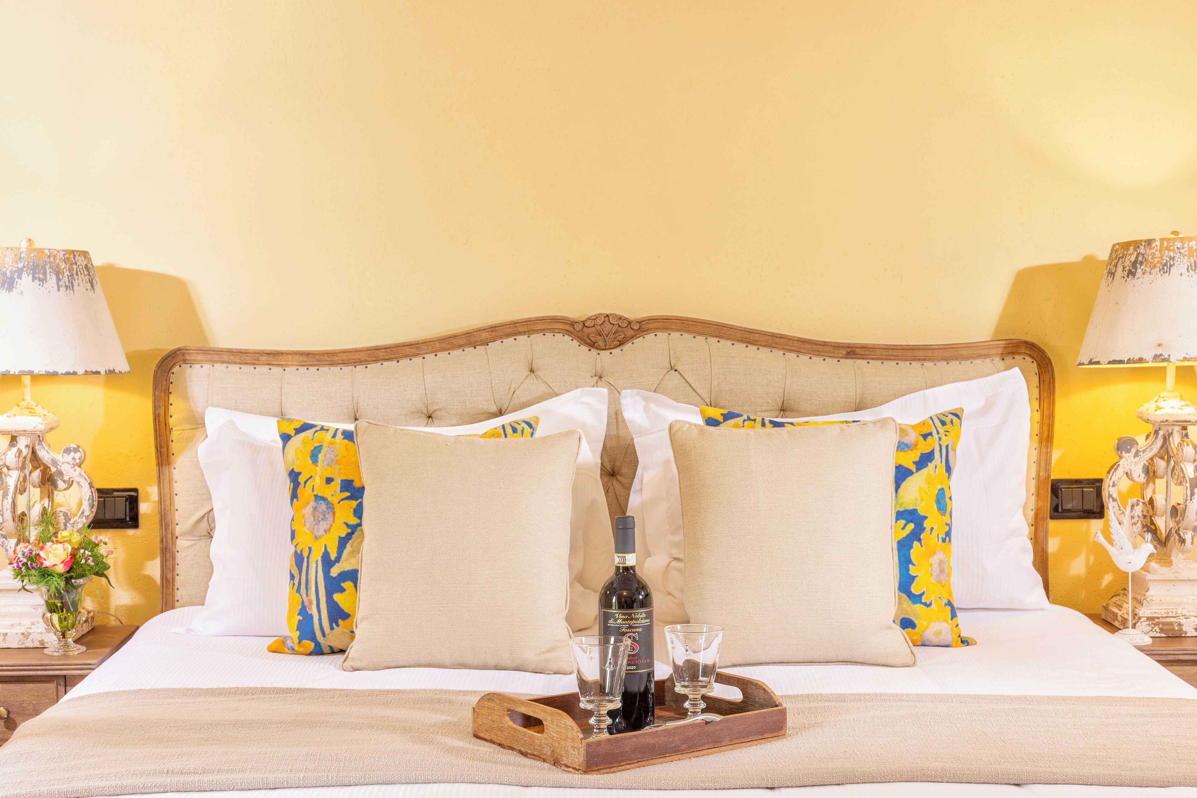 A neatly made bed with a wooden headboard, two decorative lamps, and a tray with a bottle and glasses.