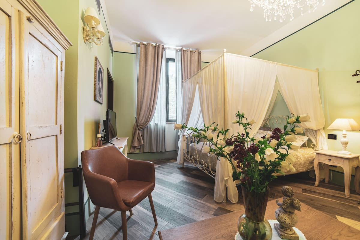 Un dormitorio con una cama con dosel, una silla, una mesita de noche y un florero con flores.