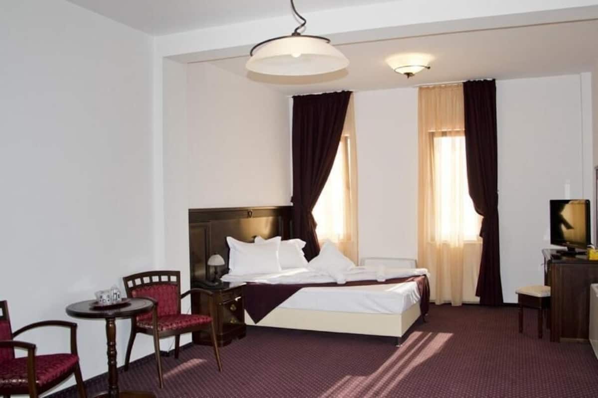 A hotel room with a bed, two chairs, a small table, a desk with a TV, and a window with curtains.