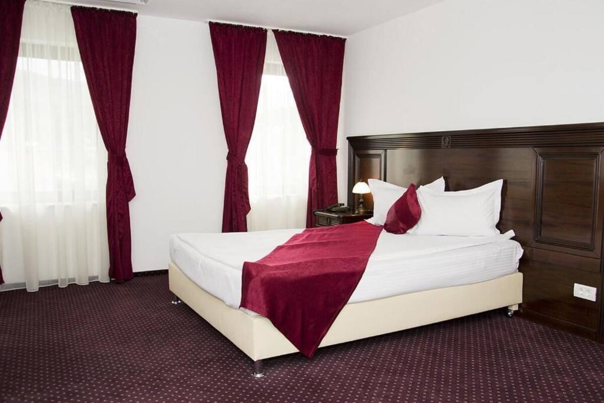A hotel room with a large bed, a wooden headboard, a nightstand with a lamp, and red curtains.