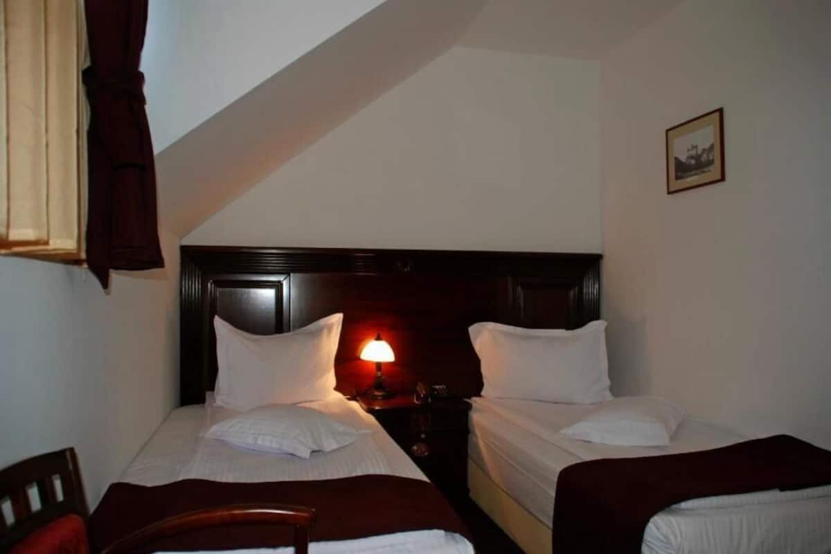 A small, dimly lit room with two single beds, a nightstand with a lamp, a chair, and a framed picture on the wall.