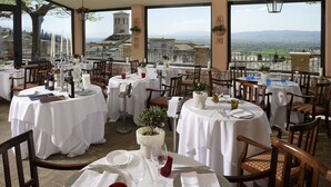 Een restaurant met meerdere tafels gedekt voor diner, met witte tafellakens en houten stoelen, en een mooi uitzicht op bergen en een toren op de achtergrond.