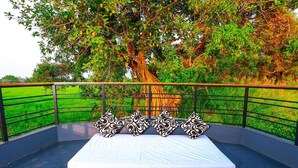 A balcony with a white mattress and four patterned pillows, surrounded by a metal railing, overlooking a green field and a large tree. - Private Houseboat (Ambalapuzha)