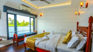 A hotel room with a large bed, a window with a view of the water, a ceiling fan, and wall-mounted lights. - Private Houseboat (Ambalapuzha)