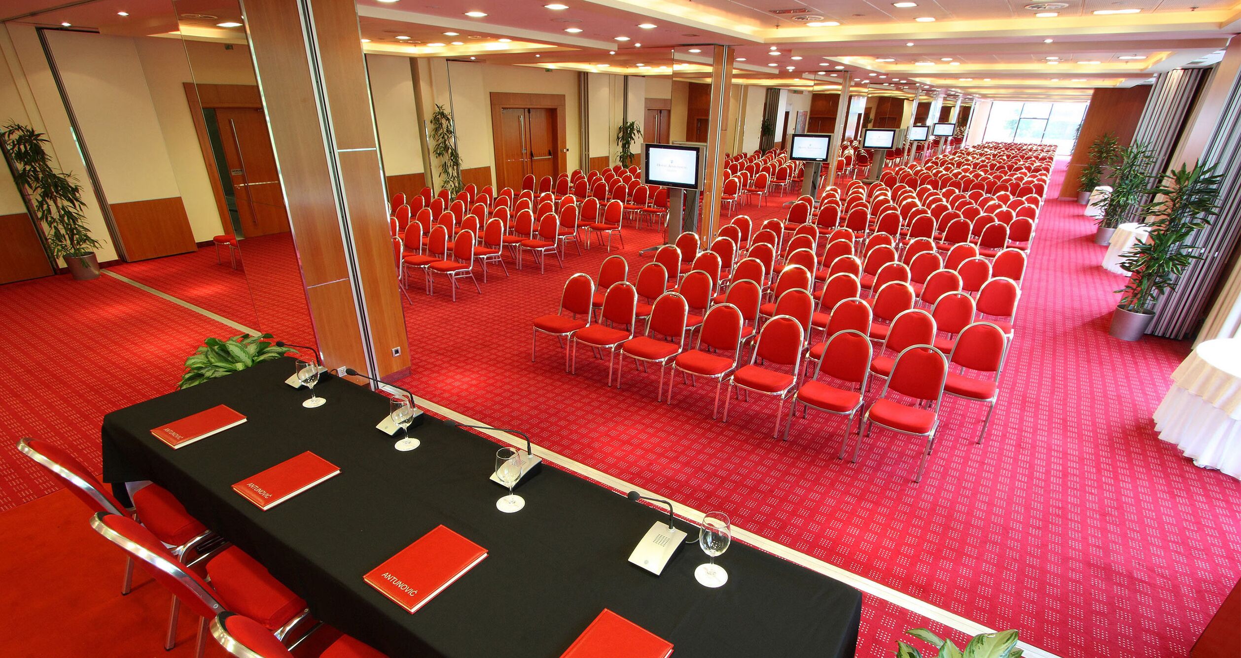 A conference room with rows of red chairs, a black table with name cards, and a red carpeted floor.