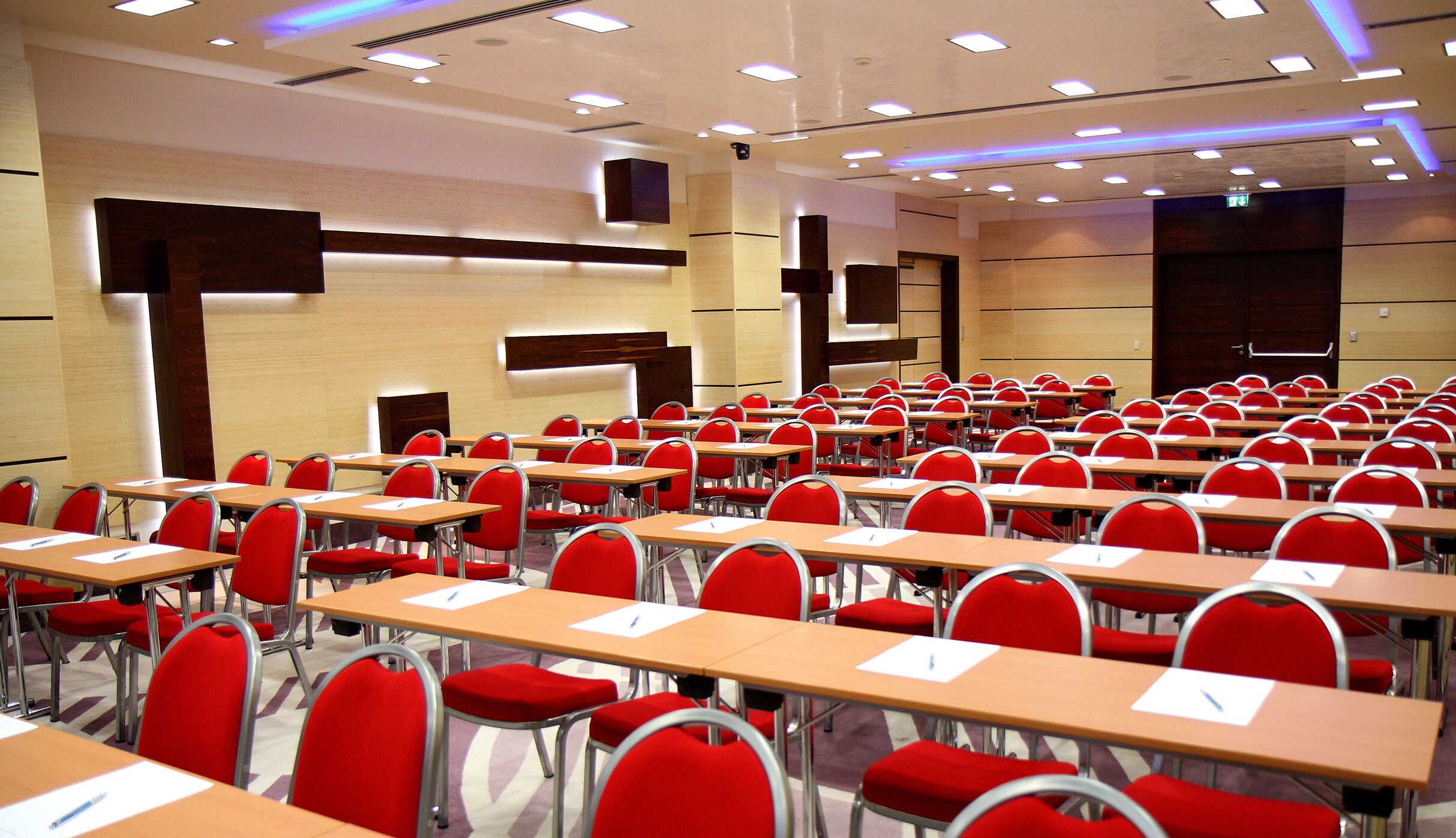 A conference room with rows of red chairs and wooden tables, equipped with overhead lighting and a modern design.