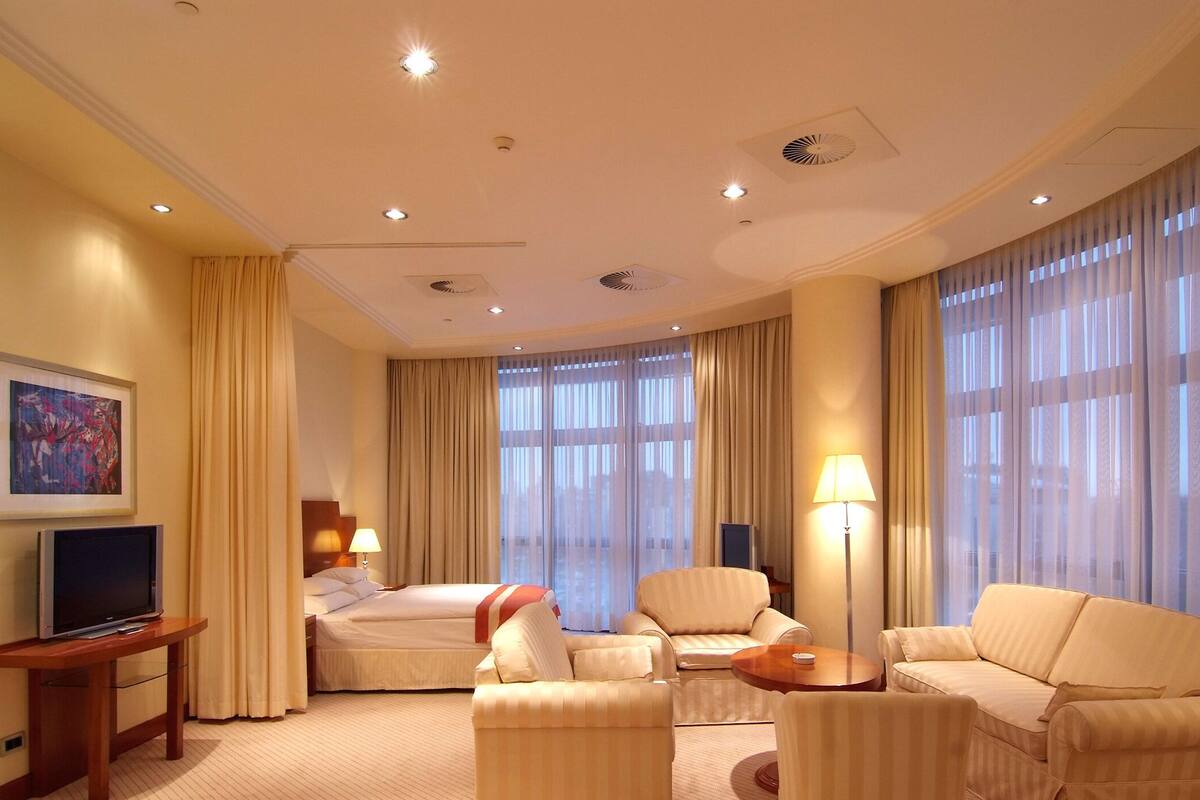 Ein Hotelzimmer mit einem Bett, zwei Sofas, einem Fernseher auf einem Schrank, einer Lampe und großen Fenstern mit Vorhängen.
