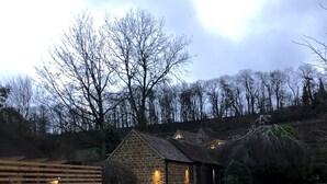 A gravel driveway leading to a stone building with a sloped roof, surrounded by trees and a wooden fence. - Wisteria Cottage - Cotswolds (Warmington)