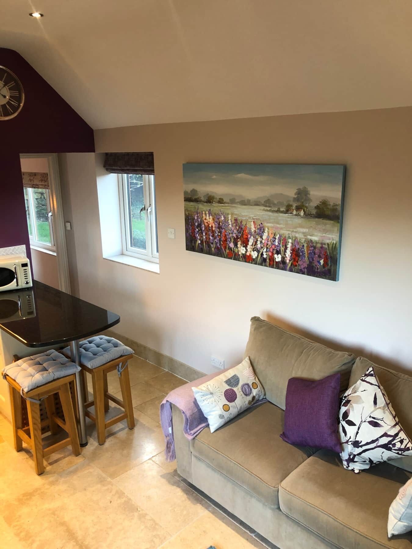 Un salon avec un canapé beige, une table à manger avec deux chaises, un tableau représentant un champ et une horloge au mur.