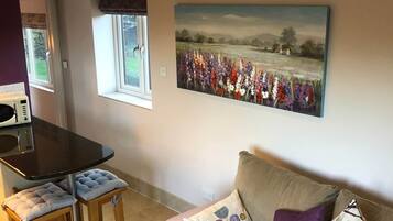 Un salon avec un canapé beige, une table à manger avec deux chaises, un tableau représentant un champ et une horloge au mur.