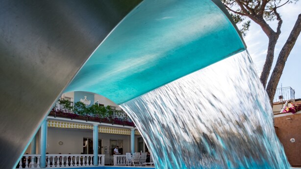 A swimming pool with a waterfall feature, surrounded by a covered area with chairs and tables. - Hotel Villa dei Fiori (Rimini)