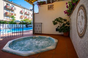 A hot tub in a courtyard with a pool in the background, surrounded by a fence and potted plants. - Hotel Villa dei Fiori (Rimini)