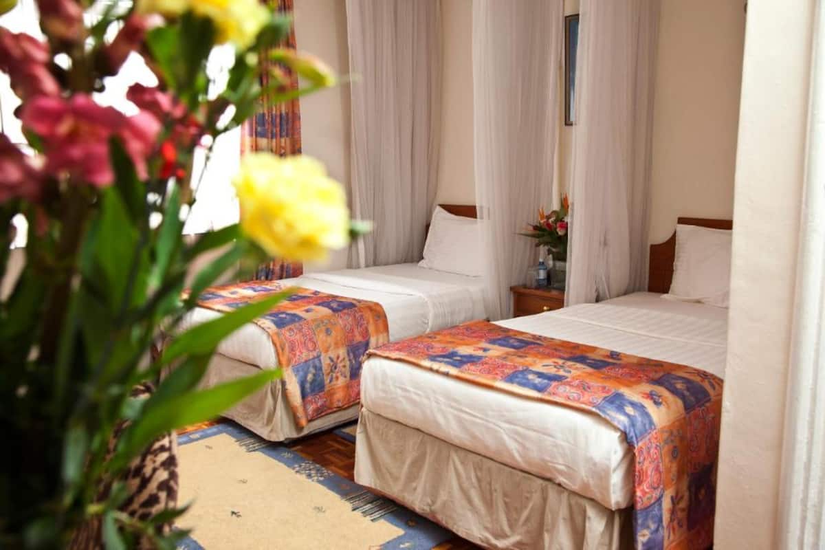 Une chambre d’hôtel avec deux lits, chacun recouvert d’une courtepointe aux couleurs vives, une table de chevet ornée d’un vase de fleurs et des rideaux en voile.