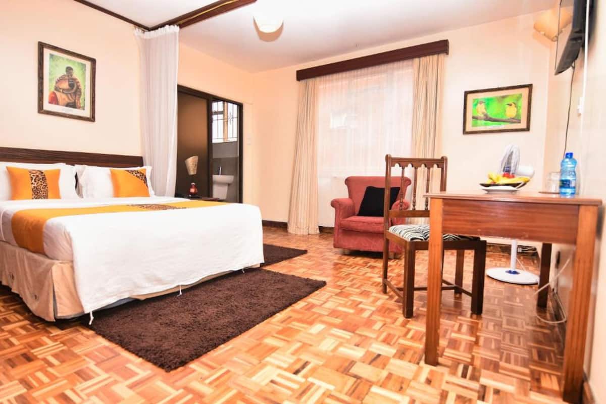 Une chambre d’hôtel comprenant un lit, un bureau, une chaise, une petite table avec un ventilateur et une fenêtre avec des rideaux.