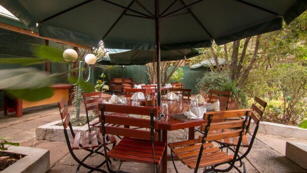 An outdoor dining area with wooden tables and chairs, covered by a large umbrella. - GraceHouse Resort (Nairobi)
