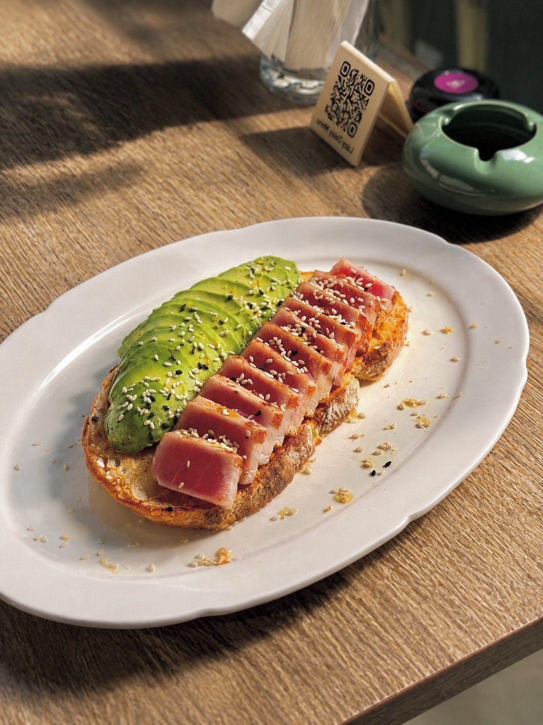 Ein Teller mit Brot, belegt mit Avocado und einer Scheibe Rohschinken, möglicherweise Thunfisch, auf einer Holztischplatte.