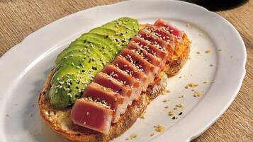 Ein Teller mit Brot, belegt mit Avocado und einer Scheibe Rohschinken, möglicherweise Thunfisch, auf einer Holztischplatte.