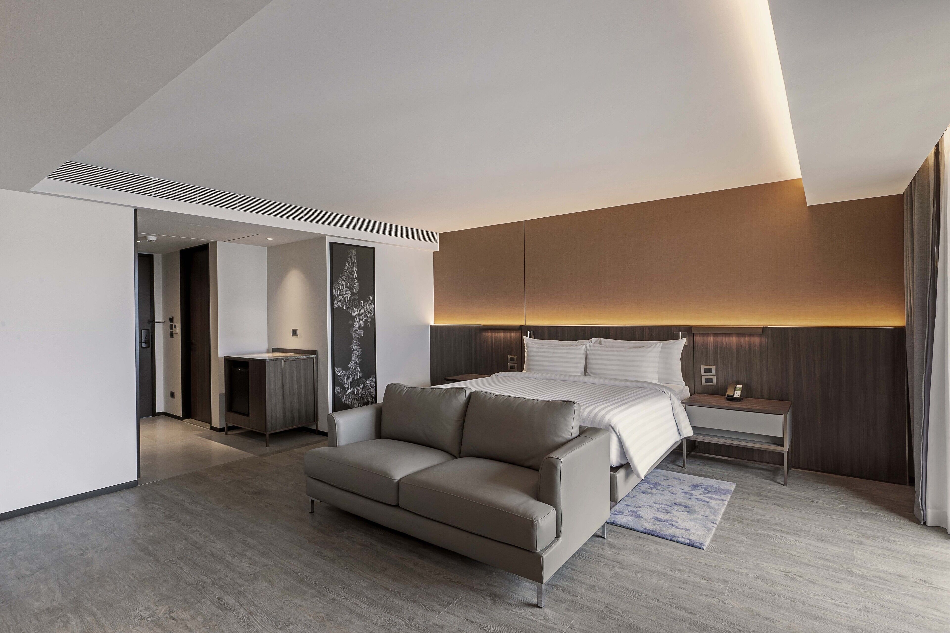 A modern hotel room with a large bed, a sofa, a bedside table, and a mirror.