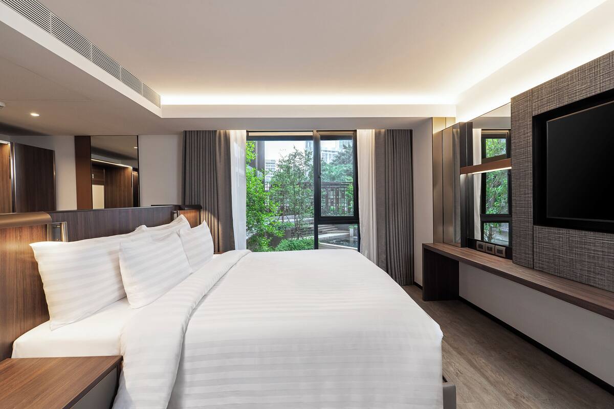 Una habitación de hotel moderna con una cama grande, un televisor y vistas a espacios verdes.