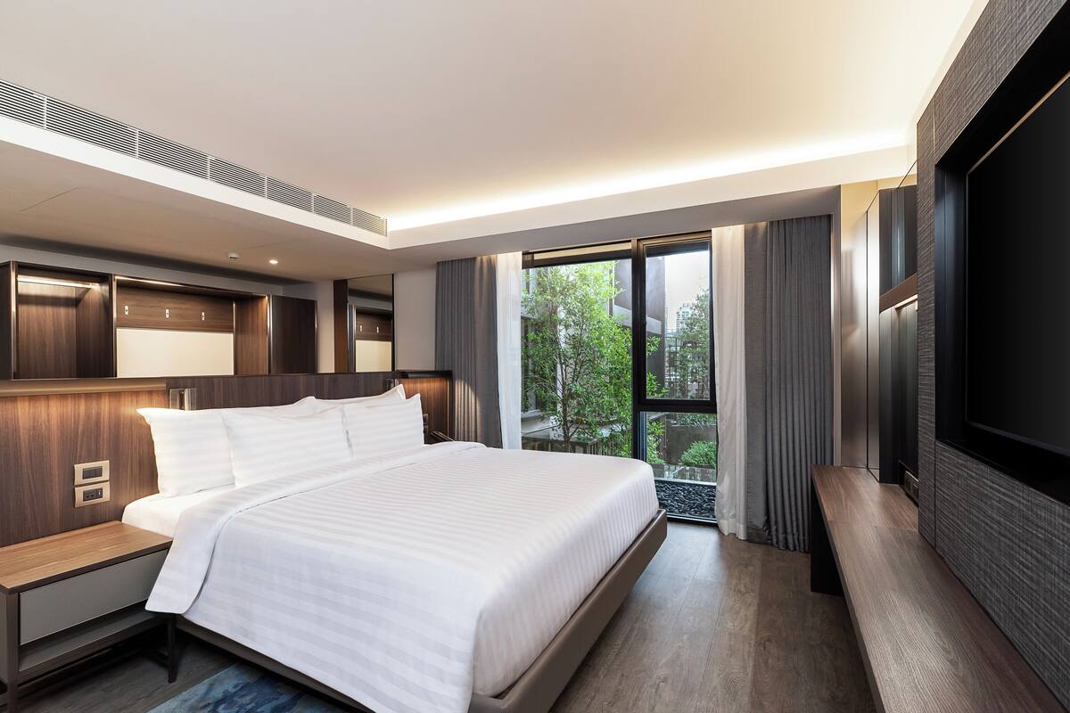 Una habitación de hotel moderna con una cama grande, un televisor de pantalla plana, un escritorio y un balcón con vistas a zonas verdes.