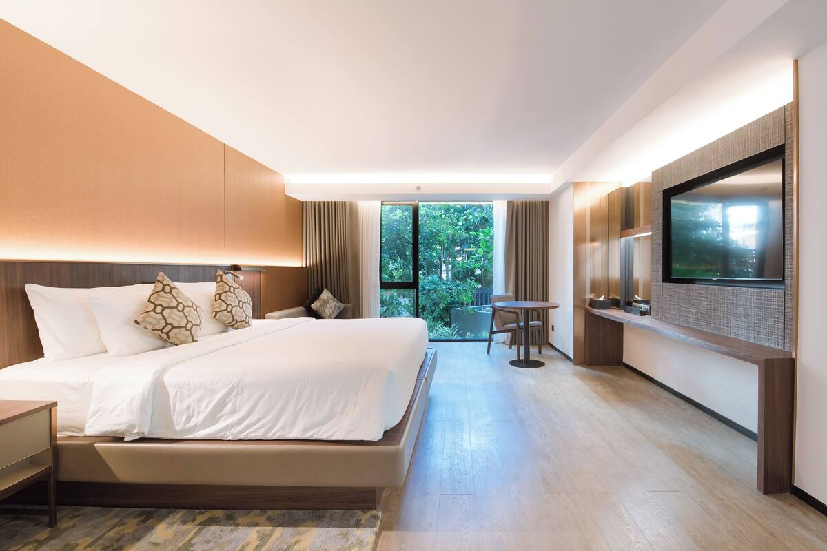 Habitación de hotel moderna con una cama grande, televisión, una mesita pequeña y vistas a la vegetación por la ventana.