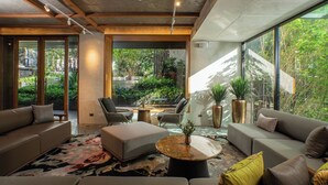 A modern living room with a sofa, ottoman, and coffee table, large windows, and indoor plants. - Gardina Asoke Hotel & Residence (Bangkok)