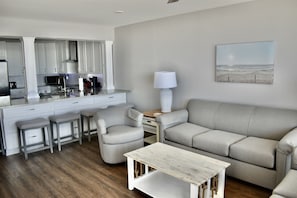 Ett modernt kök med vita skåp, en matplats med ett vitt bord och två stolar samt ett vardagsrum med en beige soffa och en lampa.