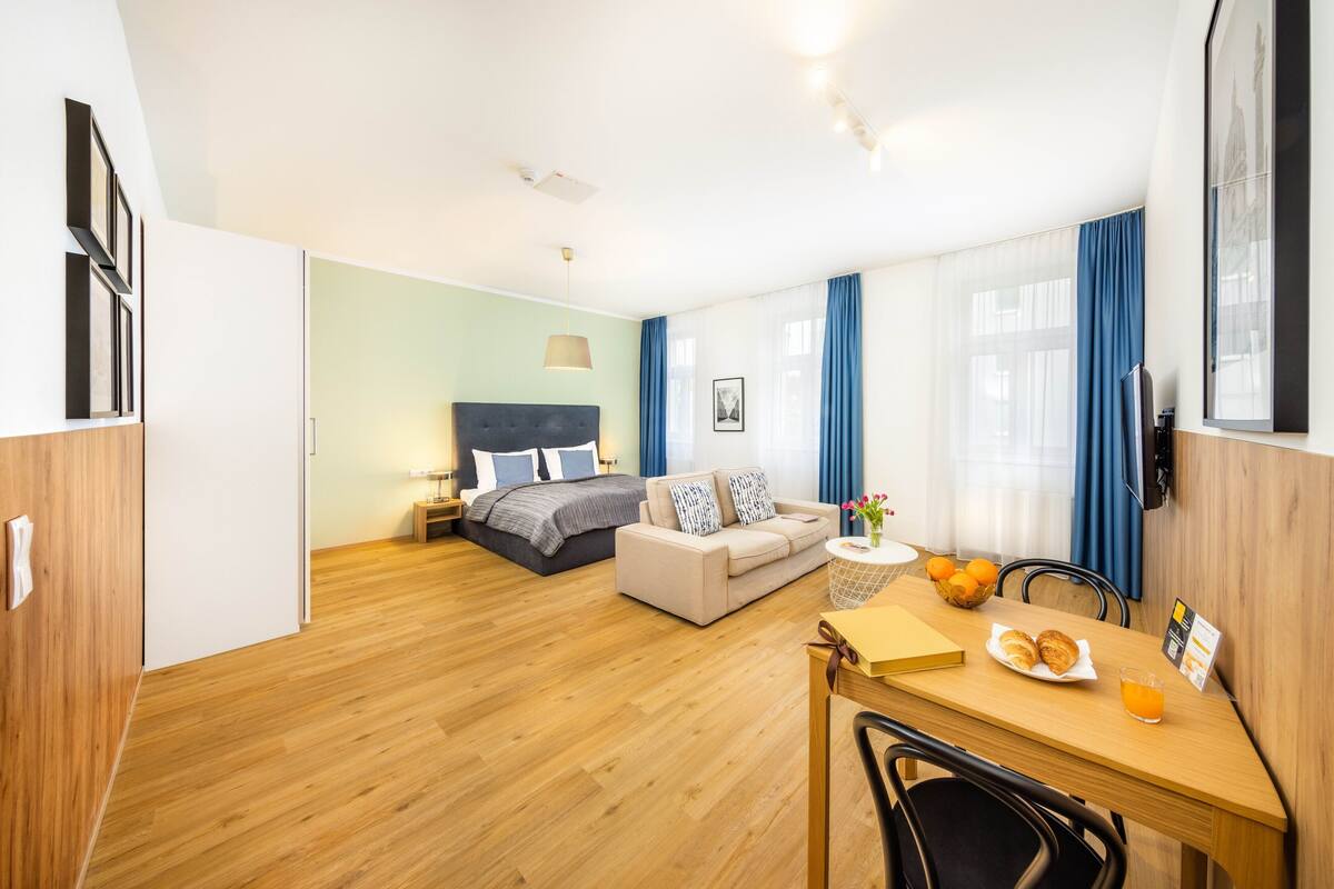 A hotel room with a bed, a sofa, a dining table with breakfast, and a large window with curtains.