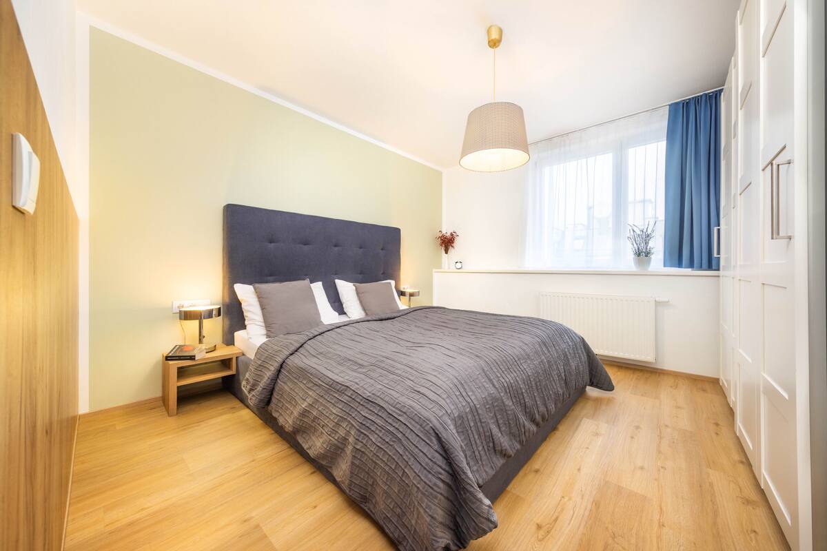 A bedroom with a large bed, a bedside table with a lamp, a window with curtains, and a radiator.