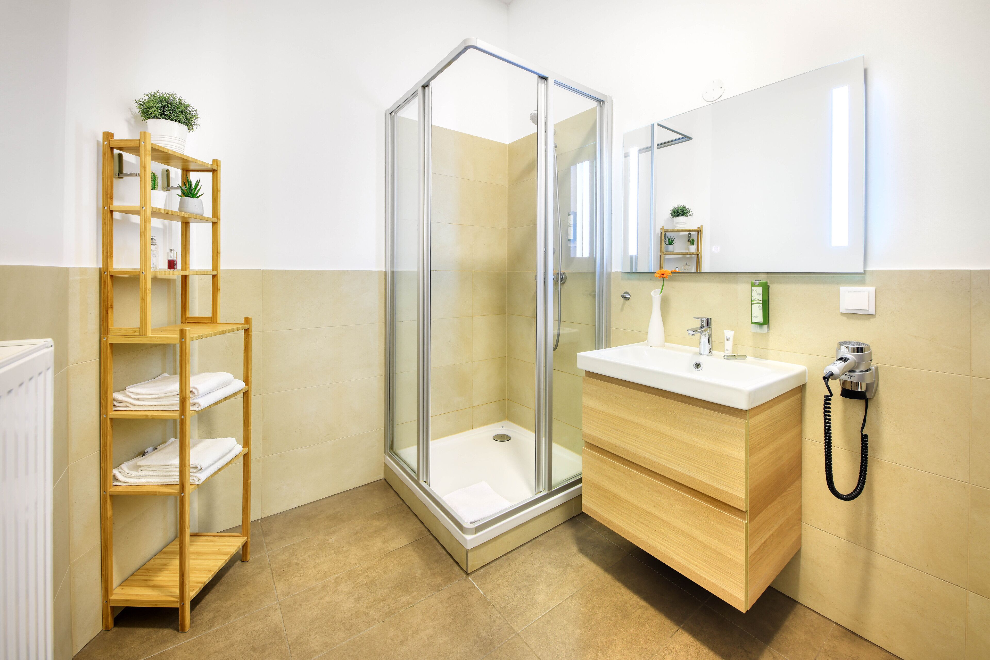 Un baño moderno con ducha de vidrio, un estante de madera con toallas, un lavabo con espejo y un secador de pelo en la pared.