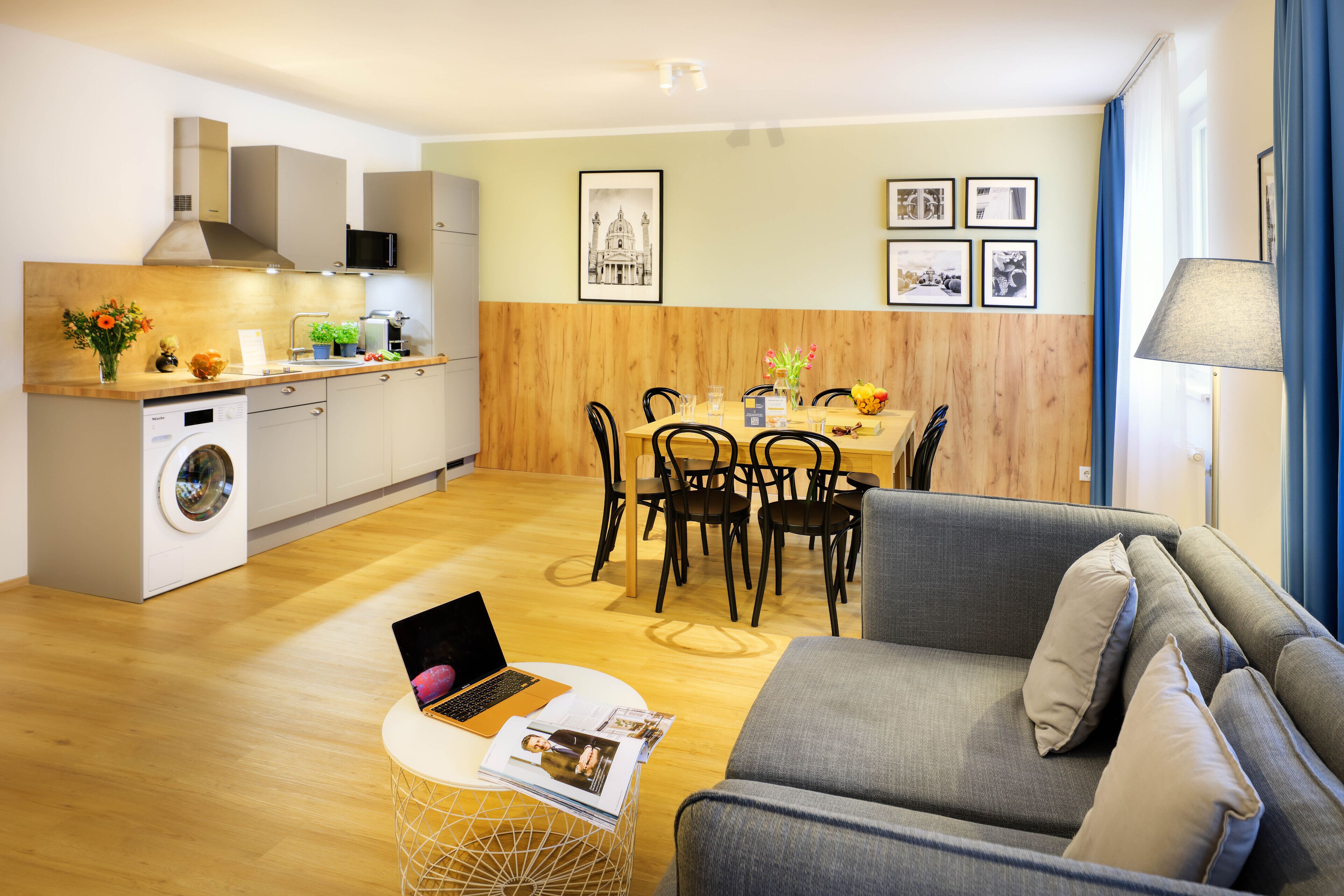 A modern kitchen with a built-in washing machine, dining area with wooden chairs, and a grey sofa.