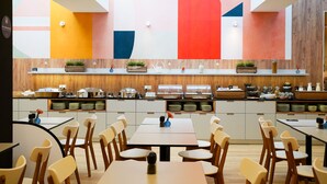 A modern dining area with multiple tables and chairs, a kitchen area with various food items and appliances, and a colorful wall design. - Jimmy's Apartments Dandelion (Vienna)