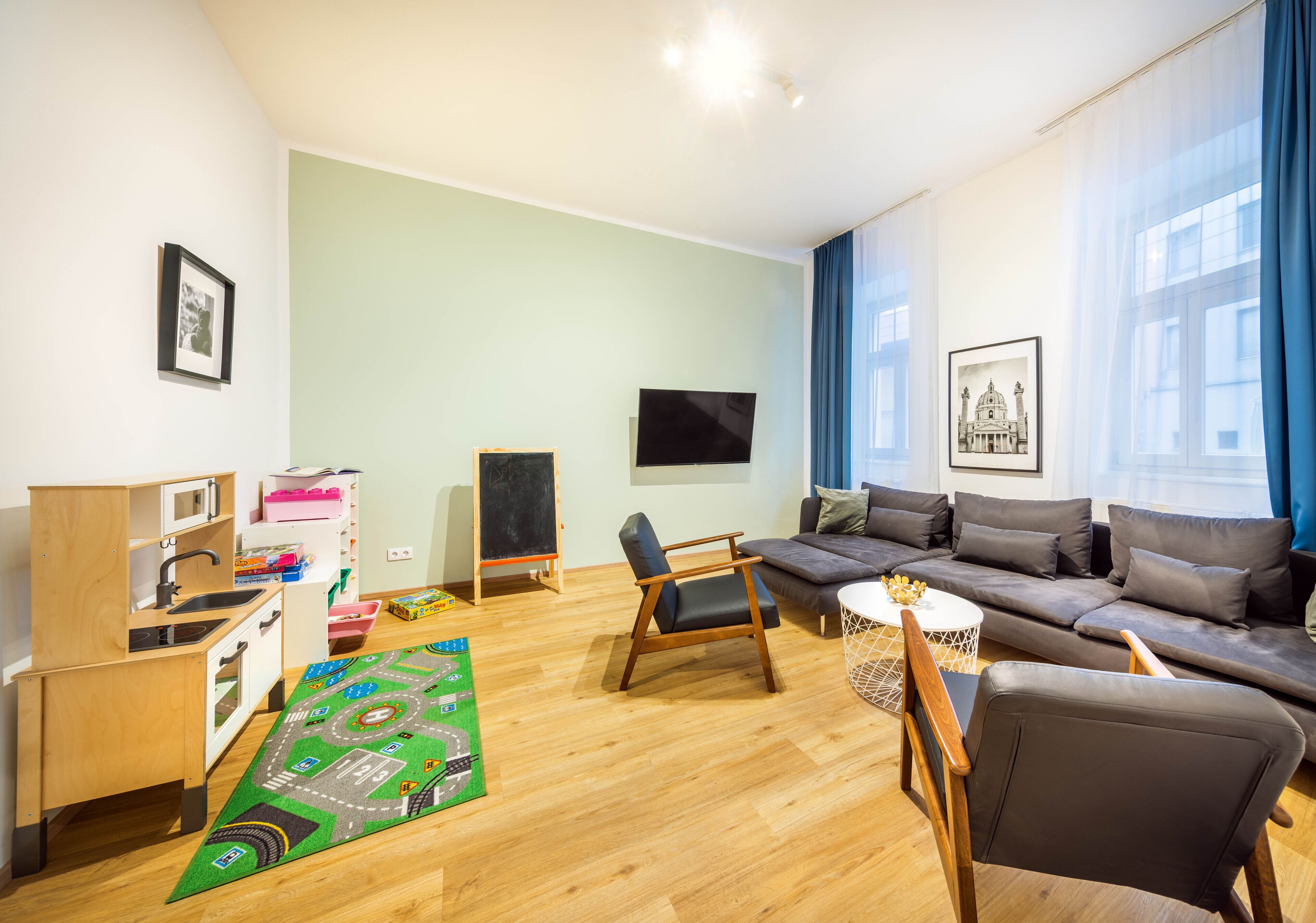 A modern living room with a sofa, chairs, a small kitchen area, and a large window with curtains.